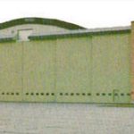 A large wood and brick quonset-type building is seen from the front. The front has six large sliding doors that are painted green. On both sides of the doors are brown brick towers anchoring the doors. On the brickwork on the right is the name "SEKINE" in white capital letters.