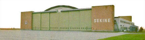 A large wood and brick quonset-type building is seen from the front. The front has six large sliding doors that are painted green. On both sides of the doors are brown brick towers anchoring the doors. On the brickwork on the right is the name "SEKINE" in white capital letters.